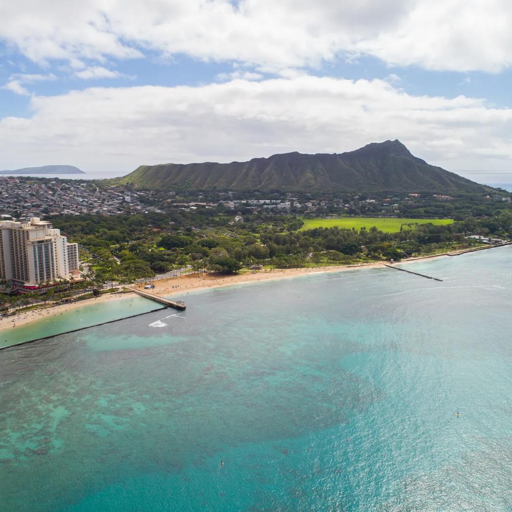 Küste mit türkisfarbenem Wasser, Sandstrand, Hochhäusern und grünem Vulkan im Hintergrund unter bewölktem Himmel.