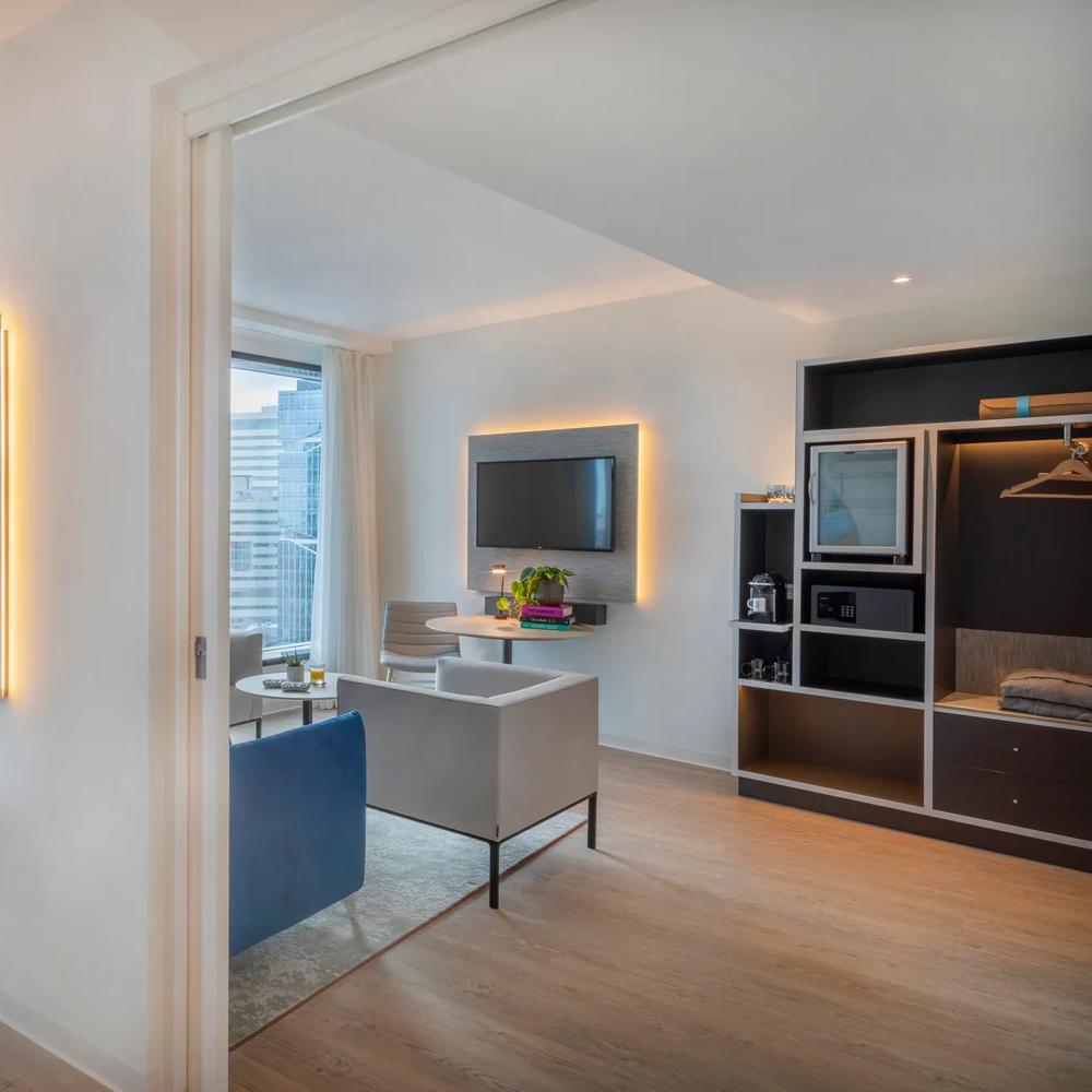 Helles Wohnzimmer mit Sofa, Tisch, Fernseher und großem Fenster mit Stadtblick. Pflanzen am Fensterbrett.