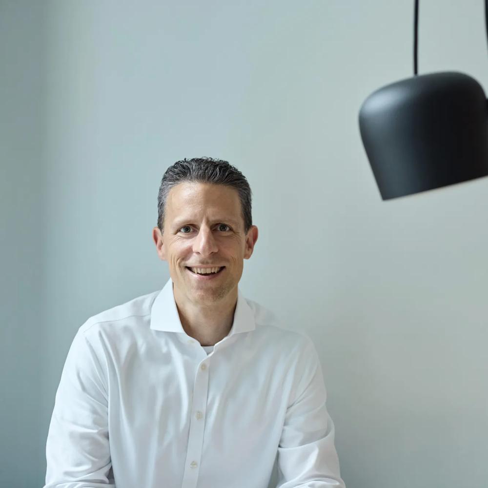 Ein Mann in weißem Hemd lächelt vor einer grauen Wand, daneben eine schwarze Hängelampe.